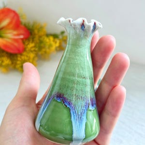 K&ouml;nnte beinhalten: Eine kleine Keramikvase mit einem ausgestellten, gewellten Rand. Der K&ouml;rper hat eine hellgr&uuml;ne Craquel&eacute;-Glasur im oberen Bereich, die in einen glatten, einfarbigen gr&uuml;nen Boden &uuml;bergeht, mit blauen und wei&szlig;en Tropfglasur-Akzenten.