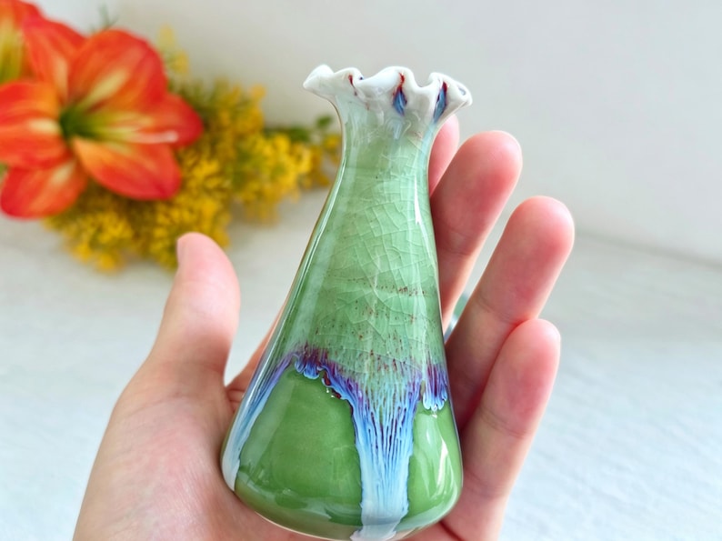 K&ouml;nnte beinhalten: Eine kleine Keramikvase mit einem ausgestellten, gewellten Rand. Der K&ouml;rper hat eine hellgr&uuml;ne Craquel&eacute;-Glasur im oberen Bereich, die in einen glatten, einfarbigen gr&uuml;nen Boden &uuml;bergeht, mit blauen und wei&szlig;en Tropfglasur-Akzenten.