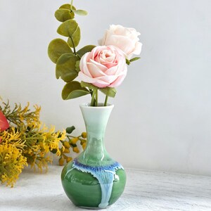 K&ouml;nnte beinhalten: Eine kleine Blumen-Vase aus Keramik mit verj&uuml;ngtem Hals und bauchigem K&ouml;rper, die eine gl&auml;nzende Glasur aufweist, welche von hellem Mintgr&uuml;n oben zu tiefem Waldgr&uuml;n unten &uuml;bergeht, akzentuiert durch ein leuchtend blaues Tropfenmuster am Hals.