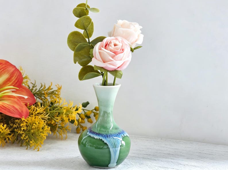 K&ouml;nnte beinhalten: Eine kleine Blumen-Vase aus Keramik mit verj&uuml;ngtem Hals und bauchigem K&ouml;rper, die eine gl&auml;nzende Glasur aufweist, welche von hellem Mintgr&uuml;n oben zu tiefem Waldgr&uuml;n unten &uuml;bergeht, akzentuiert durch ein leuchtend blaues Tropfenmuster am Hals.