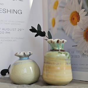 Peut inclure: Deux vases en céramique aux bords festonnés. Le vase de gauche est rond et crème, tandis que celui de droite est plus grand avec un glaçage jaune, vert et marron. Une invitation de mariage est en arrière-plan.