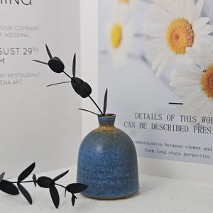 May include: A blue ceramic vase with a rounded body and a narrow neck, holding black decorative stems. The vase has a textured surface and a gradient of colors. A poster with text and daisy flowers is in the background.