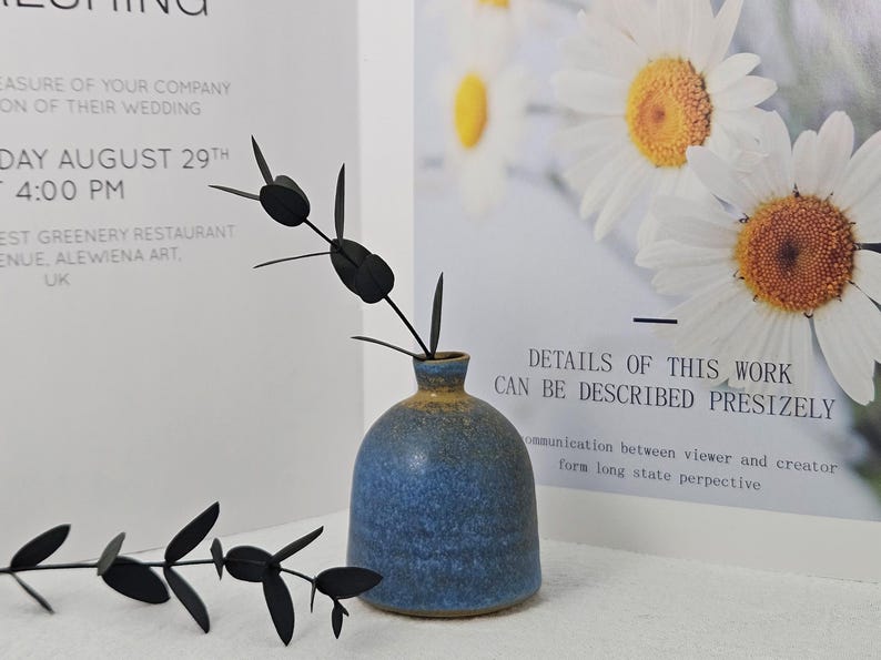 May include: A blue ceramic vase with a rounded body and a narrow neck, holding black decorative stems. The vase has a textured surface and a gradient of colors. A poster with text and daisy flowers is in the background.
