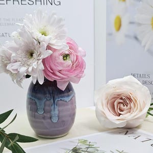 Puede incluir: Un peque&ntilde;o jarr&oacute;n de cer&aacute;mica morado y azul con una superficie texturizada contiene un ramo de flores blancas y rosas. Las flores incluyen margaritas blancas y ran&uacute;nculos rosas.