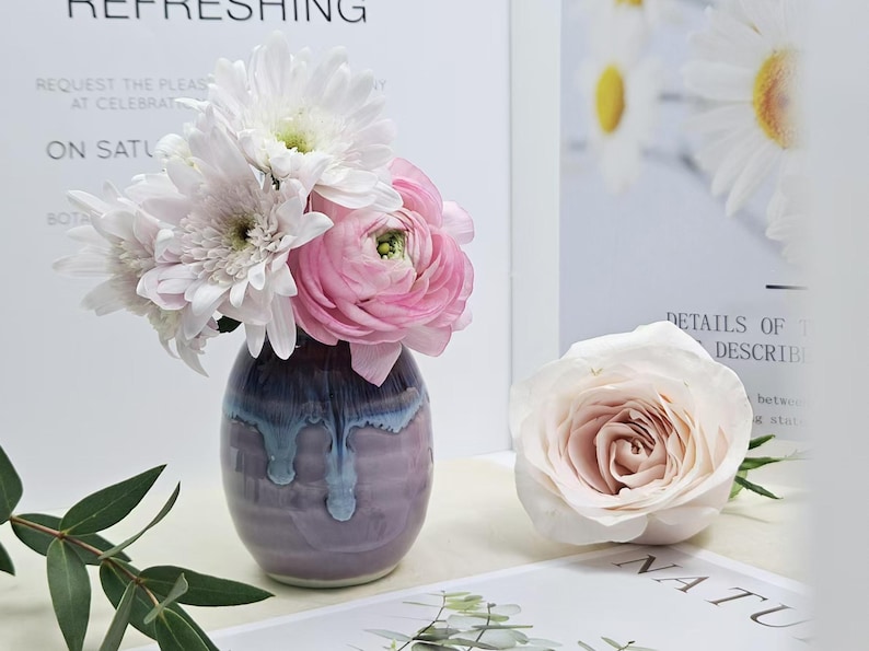 Puede incluir: Un peque&ntilde;o jarr&oacute;n de cer&aacute;mica morado y azul con una superficie texturizada contiene un ramo de flores blancas y rosas. Las flores incluyen margaritas blancas y ran&uacute;nculos rosas.