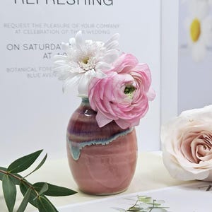 Puede incluir: Un jarr&oacute;n de cer&aacute;mica rosa con un patr&oacute;n de espiral, que contiene un ramo de flores blancas y rosas. Las flores incluyen una margarita blanca y ran&uacute;nculos rosas.