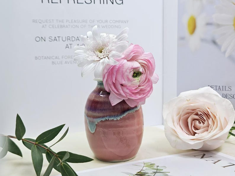 Puede incluir: Un jarr&oacute;n de cer&aacute;mica rosa con un patr&oacute;n de espiral, que contiene un ramo de flores blancas y rosas. Las flores incluyen una margarita blanca y ran&uacute;nculos rosas.