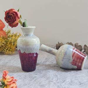 May include: Two ceramic vases with a gradient color scheme. One vase is upright, holding a dried rose. The other vase is lying on its side. The vases transition from white to blue to a deep burgundy.