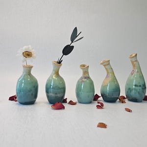 May include: Five small ceramic vases in shades of green and brown with a white flower in one of the vases. The vases are arranged in a row on a white surface.