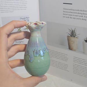 K&ouml;nnte beinhalten: Eine kleine, handgefertigte Keramikvase mit einer einzigartigen Form und einem gl&auml;nzenden, strukturierten Finish. Die Vase hat eine Mischung aus gr&uuml;nen, blauen und wei&szlig;en Farben mit einem ger&uuml;schten Oberteil. Die Vase wird in einer Hand gehalten, mit einem Buch im Hintergrund.