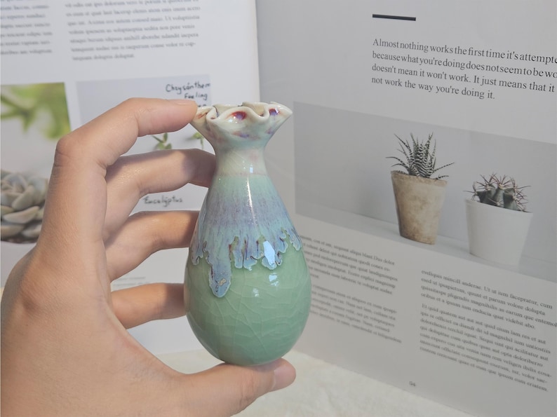 K&ouml;nnte beinhalten: Eine kleine, handgefertigte Keramikvase mit einer einzigartigen Form und einem gl&auml;nzenden, strukturierten Finish. Die Vase hat eine Mischung aus gr&uuml;nen, blauen und wei&szlig;en Farben mit einem ger&uuml;schten Oberteil. Die Vase wird in einer Hand gehalten, mit einem Buch im Hintergrund.