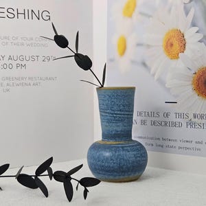 May include: A small, blue ceramic vase with a bulbous base and a textured surface. The vase has a yellow rim and holds black, leaf-like stems. The background includes text and a floral print.