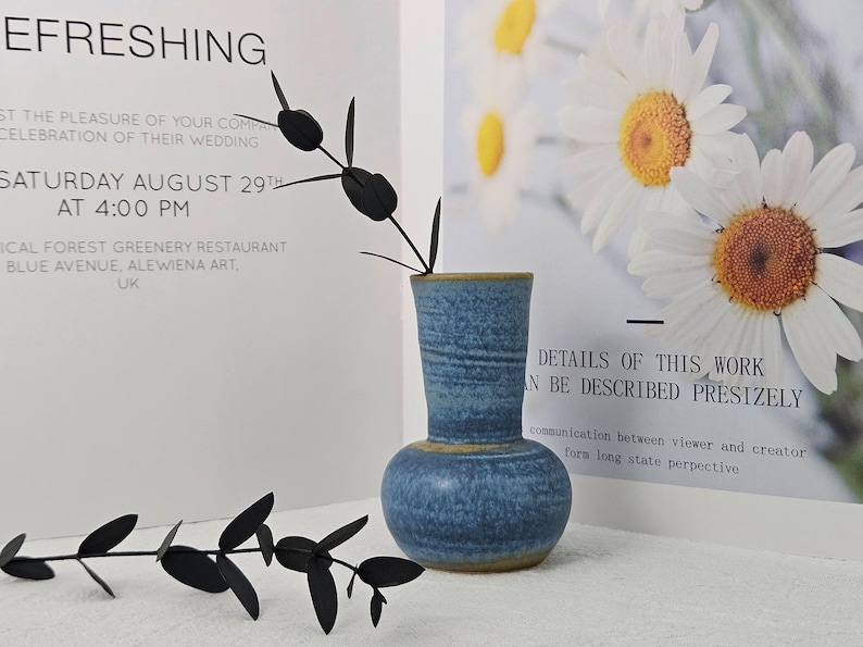May include: A small, blue ceramic vase with a bulbous base and a textured surface. The vase has a yellow rim and holds black, leaf-like stems. The background includes text and a floral print.