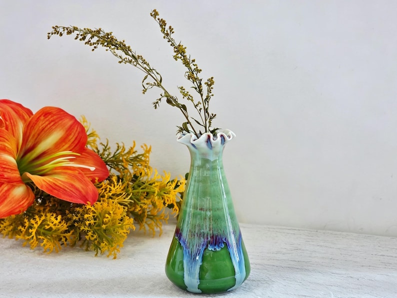K&ouml;nnte beinhalten: Eine Keramikvase mit einem ausgestellten, ger&uuml;schten Rand und einem konischen K&ouml;rper. Die Glasur zeigt einen Verlauf von Hellgr&uuml;n oben, der in ein dunkelviolettes und blaues Tropfmuster auf einer leuchtend gr&uuml;nen Basis &uuml;bergeht, mit braunen Stielen.