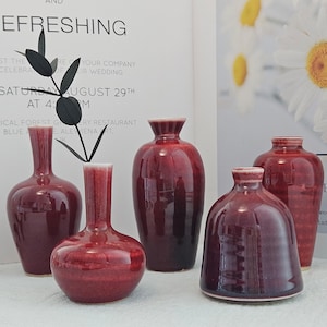 Peut inclure: Ensemble de cinq vases en céramique bordeaux à bords blancs. Les vases, de formes et tailles variées, présentent des surfaces brillantes. En arrière-plan, une affiche avec le mot "REFRESHING" et des motifs floraux.