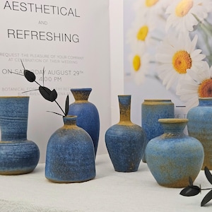 May include: A selection of blue ceramic vases with a textured finish and diverse shapes. The vases are displayed on a white surface, with text reading "AESTHETICAL AND REFRESHING". Daisies are visible in the background.