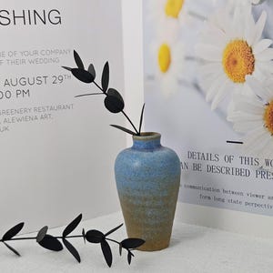 May include: A small, blue and tan ceramic vase holds a black, leaf-shaped decorative stem. The vase is set against a backdrop of white paper with text, and a floral print with daisies.