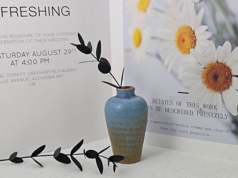 May include: A small, blue and tan ceramic vase holds a black, leaf-shaped decorative stem. The vase is set against a backdrop of white paper with text, and a floral print with daisies.