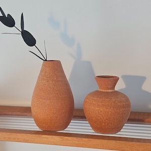 May include: Two terracotta vases of different sizes sit on a wooden shelf. The taller vase holds a black, stylized floral arrangement. The vases have a textured surface and a warm, earthy tone, creating a minimalist aesthetic.