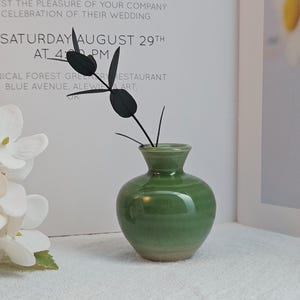 Peut inclure: Un petit vase rond en céramique verte à col étroit, contenant une plante sombre et stylisée. Le vase est posé sur une surface blanche, avec une invitation de mariage en arrière-plan. L'invitation indique "ON SATURDAY AUGUST 29TH AT 4 PM."