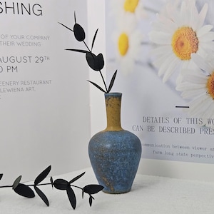 May include: A blue and yellow ceramic vase holds black decorative stems. The vase has a rounded body and a narrow neck. The background includes text about a wedding and a floral print.