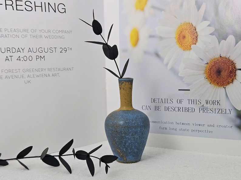May include: A blue and yellow ceramic vase holds black decorative stems. The vase has a rounded body and a narrow neck. The background includes text about a wedding and a floral print.