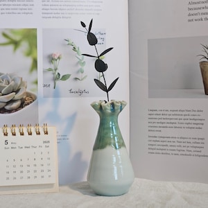 K&ouml;nnte beinhalten: Eine Keramikvase mit wei&szlig;em Sockel und gr&uuml;nem Oberteil, die eine schwarze dekorative Pflanze enth&auml;lt. Ein kleiner Kalender zeigt das Datum 5. Mai 2025. Der Hintergrund umfasst Seiten aus einem Buch mit Pflanzenbildern.