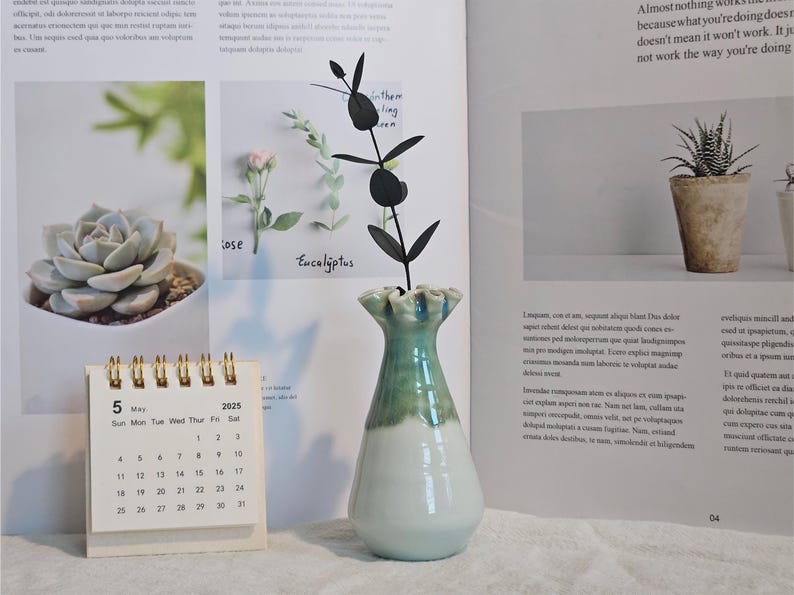 K&ouml;nnte beinhalten: Eine Keramikvase mit wei&szlig;em Sockel und gr&uuml;nem Oberteil, die eine schwarze dekorative Pflanze enth&auml;lt. Ein kleiner Kalender zeigt das Datum 5. Mai 2025. Der Hintergrund umfasst Seiten aus einem Buch mit Pflanzenbildern.