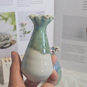 K&ouml;nnte beinhalten: Kleine, handgefertigte Keramikvase mit ger&uuml;schtem Rand. Die Vase ist am Boden wei&szlig; und geht in ein helles Gr&uuml;n mit Craquel&eacute;-Glasur &uuml;ber. Die Vase wird in einer Hand gehalten.
