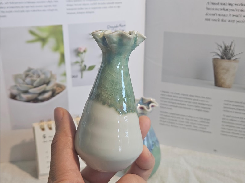 K&ouml;nnte beinhalten: Kleine, handgefertigte Keramikvase mit ger&uuml;schtem Rand. Die Vase ist am Boden wei&szlig; und geht in ein helles Gr&uuml;n mit Craquel&eacute;-Glasur &uuml;ber. Die Vase wird in einer Hand gehalten.