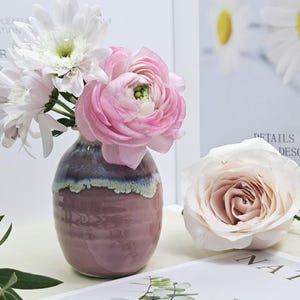 Puede incluir: Un peque&ntilde;o jarr&oacute;n de cer&aacute;mica rosa y morado con un borde blanco, lleno de flores blancas y rosas. El jarr&oacute;n est&aacute; sobre una superficie blanca.