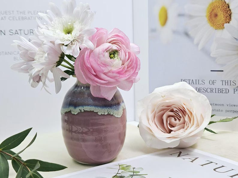 Puede incluir: Un peque&ntilde;o jarr&oacute;n de cer&aacute;mica rosa y morado con un borde blanco, lleno de flores blancas y rosas. El jarr&oacute;n est&aacute; sobre una superficie blanca.