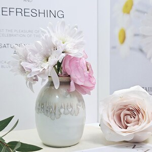 Puede incluir: Un jarr&oacute;n de cer&aacute;mica blanca con un esmalte texturizado, lleno de flores blancas y rosas. El jarr&oacute;n est&aacute; sobre una superficie blanca.