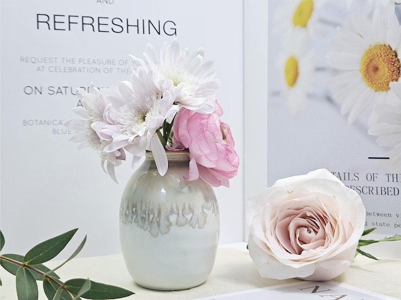 Puede incluir: Un jarr&oacute;n de cer&aacute;mica blanca con un esmalte texturizado, lleno de flores blancas y rosas. El jarr&oacute;n est&aacute; sobre una superficie blanca.