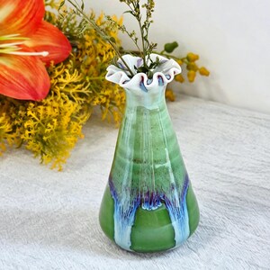 K&ouml;nnte beinhalten: Eine Keramik-Blumenvase mit einem ausgestellten, ger&uuml;schten Rand. Der K&ouml;rper ist mit einer gl&auml;nzenden gr&uuml;nen Glasur &uuml;berzogen, wobei vertikale Tropfmuster in dunklen Violett- und Hellblaut&ouml;nen an den Seiten herablaufen.