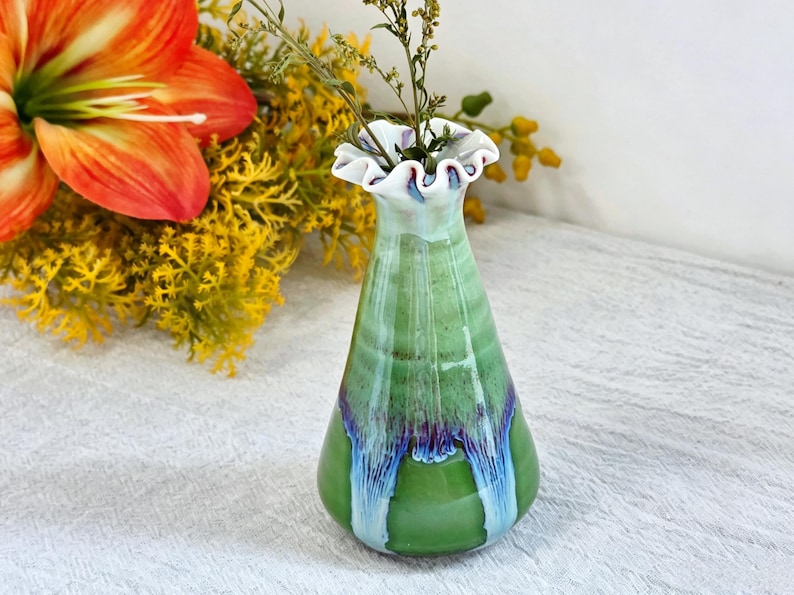 K&ouml;nnte beinhalten: Eine Keramik-Blumenvase mit einem ausgestellten, ger&uuml;schten Rand. Der K&ouml;rper ist mit einer gl&auml;nzenden gr&uuml;nen Glasur &uuml;berzogen, wobei vertikale Tropfmuster in dunklen Violett- und Hellblaut&ouml;nen an den Seiten herablaufen.