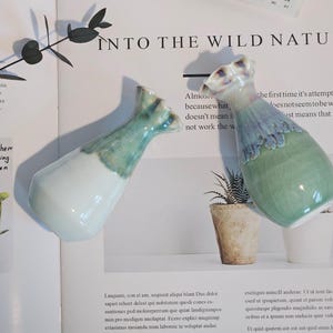 K&ouml;nnte beinhalten: Zwei Keramikvasen mit einer einzigartigen Form und einem zweifarbigen Farbschema. Die Vasen sind an der Basis wei&szlig; und gehen oben in ein t&uuml;rkisgr&uuml;n &uuml;ber. Der Text "INTO THE WILD NATURE" ist sichtbar.