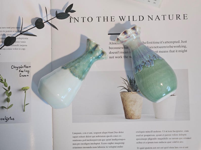 K&ouml;nnte beinhalten: Zwei Keramikvasen mit einer einzigartigen Form und einem zweifarbigen Farbschema. Die Vasen sind an der Basis wei&szlig; und gehen oben in ein t&uuml;rkisgr&uuml;n &uuml;ber. Der Text "INTO THE WILD NATURE" ist sichtbar.