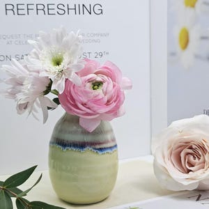 Puede incluir: Un peque&ntilde;o jarr&oacute;n de cer&aacute;mica verde claro con un dise&ntilde;o de rayas azules y moradas. El jarr&oacute;n est&aacute; lleno de flores blancas y rosas.