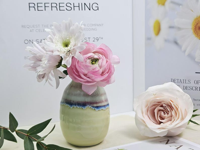 Puede incluir: Un peque&ntilde;o jarr&oacute;n de cer&aacute;mica verde claro con un dise&ntilde;o de rayas azules y moradas. El jarr&oacute;n est&aacute; lleno de flores blancas y rosas.