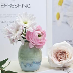 Puede incluir: Un peque&ntilde;o jarr&oacute;n de cer&aacute;mica verde claro y azul con un dise&ntilde;o ondulado contiene un ramo de flores blancas y rosas. Las flores incluyen margaritas blancas y ran&uacute;nculos rosas.