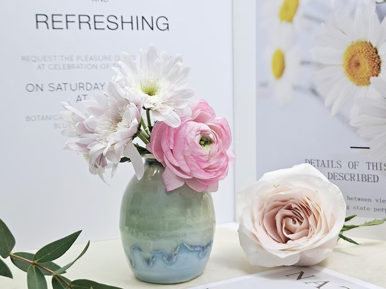 Puede incluir: Un peque&ntilde;o jarr&oacute;n de cer&aacute;mica verde claro y azul con un dise&ntilde;o ondulado contiene un ramo de flores blancas y rosas. Las flores incluyen margaritas blancas y ran&uacute;nculos rosas.