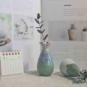 K&ouml;nnte beinhalten: Eine gr&uuml;n-blaue Keramikvase mit ger&uuml;schtem Rand, die einen dunklen Pflanzenstiel h&auml;lt, steht neben einer umgefallenen Vase. Ein kleiner Kalender zeigt das Datum 5. Mai 2025. Das Bild enth&auml;lt eine Sukkulente und kleine Topfpflanzen.
