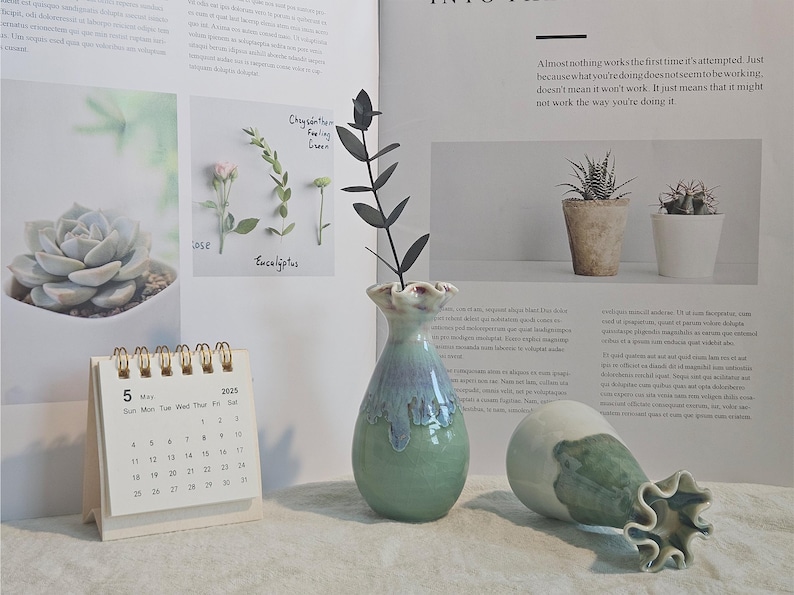 K&ouml;nnte beinhalten: Eine gr&uuml;n-blaue Keramikvase mit ger&uuml;schtem Rand, die einen dunklen Pflanzenstiel h&auml;lt, steht neben einer umgefallenen Vase. Ein kleiner Kalender zeigt das Datum 5. Mai 2025. Das Bild enth&auml;lt eine Sukkulente und kleine Topfpflanzen.