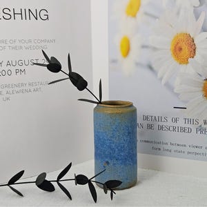 May include: A blue and tan ceramic vase with a dark green plant stem. The vase is cylindrical and has a textured surface. The background includes text about a wedding and a floral print.