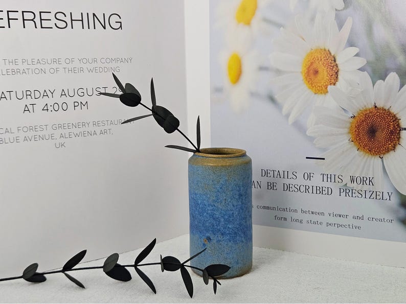 May include: A blue and tan ceramic vase with a dark green plant stem. The vase is cylindrical and has a textured surface. The background includes text about a wedding and a floral print.