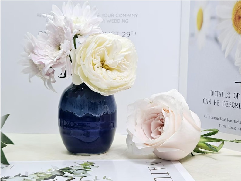 Puede incluir: Un jarr&oacute;n de cer&aacute;mica azul con un glaseado blanco en el borde superior contiene un ramo de rosas blancas y rosas y crisantemos blancos.