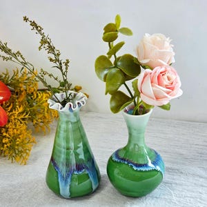 K&ouml;nnte beinhalten: Zwei kleine Keramikvasen mit gl&auml;nzender gr&uuml;ner Glasur und blau-violetten Tropf-Akzenten. Die linke Vase hat einen ausgestellten, ger&uuml;schten Rand, w&auml;hrend die rechte Vase einen runden K&ouml;rper und einen schmalen Hals hat. Beide enthalten Kunstblumen.