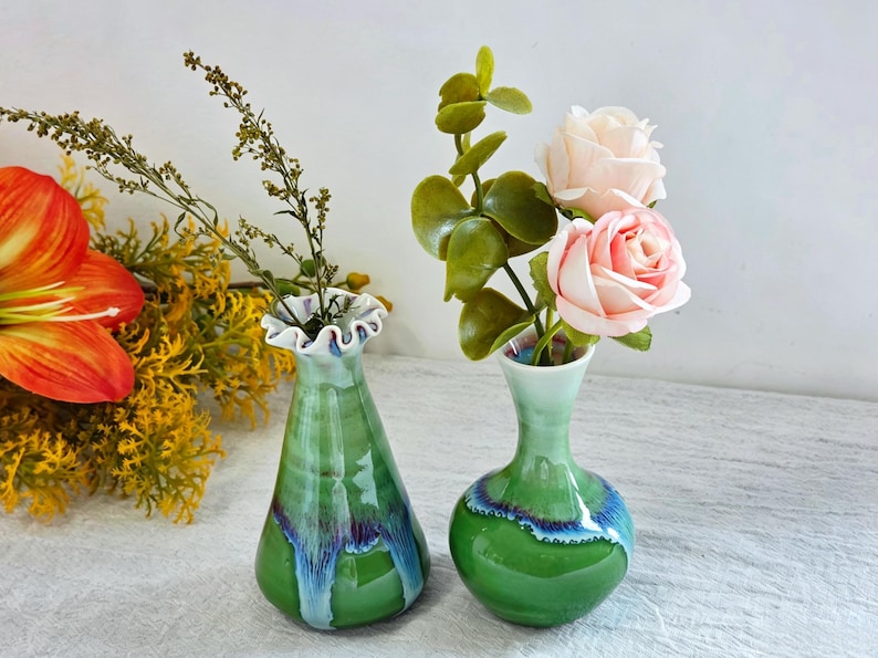 K&ouml;nnte beinhalten: Zwei kleine Keramikvasen mit gl&auml;nzender gr&uuml;ner Glasur und blau-violetten Tropf-Akzenten. Die linke Vase hat einen ausgestellten, ger&uuml;schten Rand, w&auml;hrend die rechte Vase einen runden K&ouml;rper und einen schmalen Hals hat. Beide enthalten Kunstblumen.