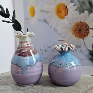 May include: Two ceramic vases with ruffled rims. One vase is a teardrop shape with a lavender base, light blue middle, and maroon top. The other vase is round with a lavender base and a light blue top. The background includes a daisy print.